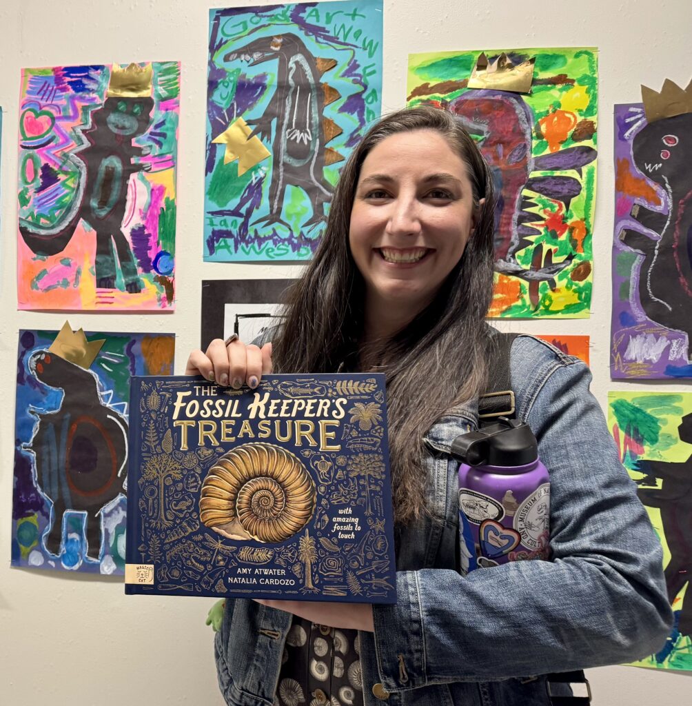 Amy Atwater holds her book in front of a wall of children's art.